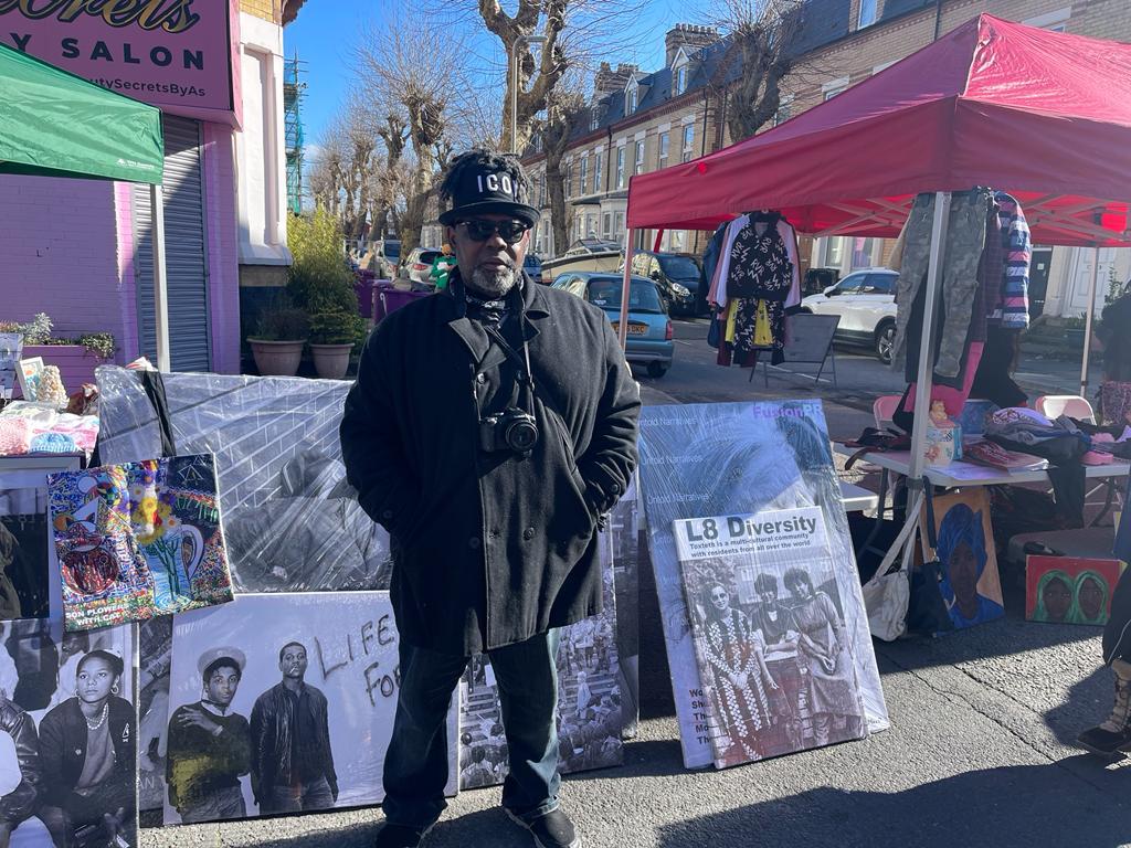 A man standing in front of artwork being sold at a market stall.